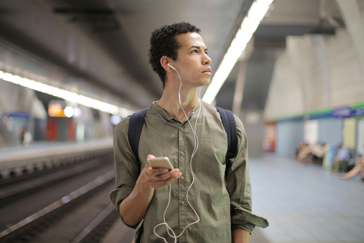 Young ethnic man in earbuds listening to music while waiting for transport at contemporary subway station