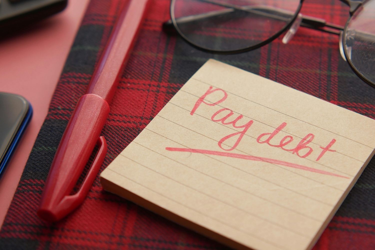 Close-up of a note reading 'Pay debt' next to a red pen on a plaid fabric, emphasizing financial reminders.